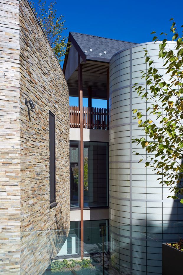 A close architectural exterior view of a contemporary multi-level building or courtyard. A curved wall clad in thin, stacked natural stone is juxtaposed with a large cylindrical form finished in pale horizontal metal or panel bands. Between them is a glazed circulation or courtyard zone with dark framing, timber vertical screening, a pitched dark roof, and a glass balustrade. The image is brightly lit against a clear blue sky, with some planting in the foreground.