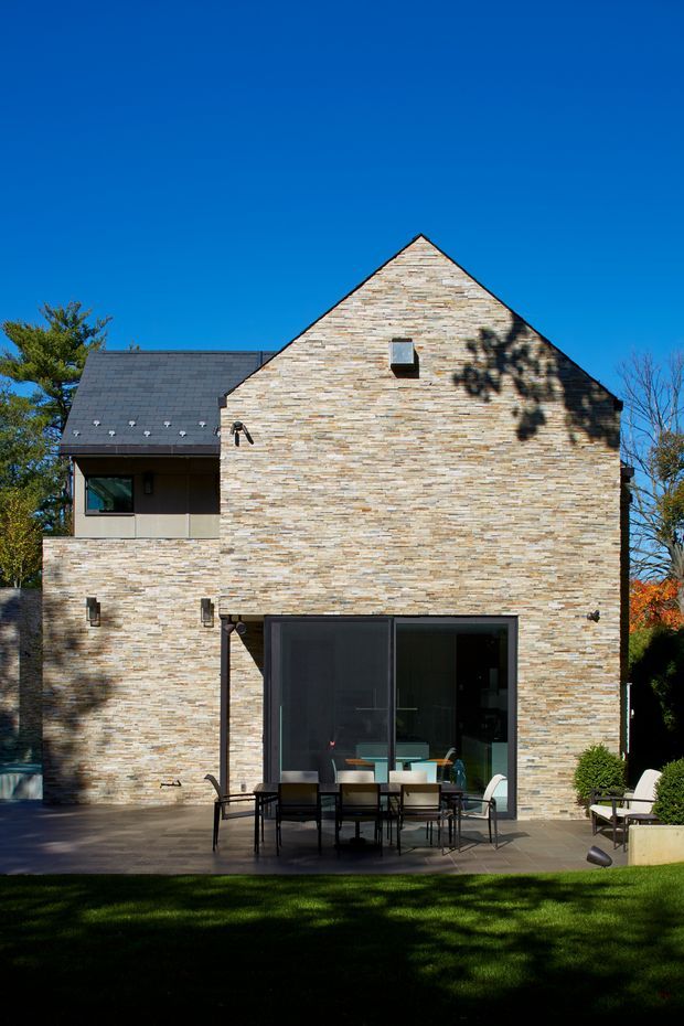 Exterior rear elevation of a contemporary multi-level residence with a steep gable roof and extensive pale stacked-stone cladding. Large dark-framed glazed doors open onto a paved outdoor dining terrace, with lawn and landscaped planting in the foreground. A slate-like roof, restrained detailing, and strong geometric massing are visible under a clear blue sky.