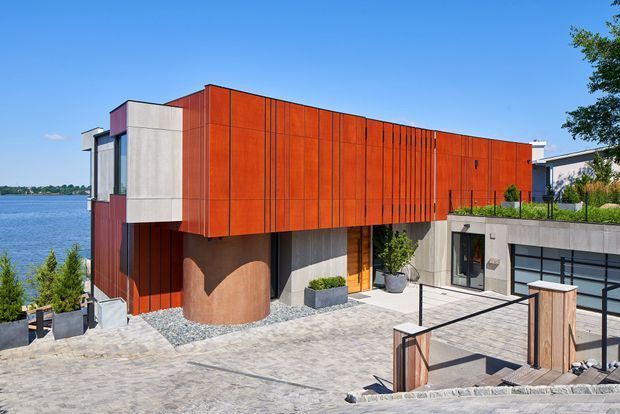 A contemporary multi-level waterfront residence with a bold cantilevered upper volume clad in vivid rust-red vertical panels. The home combines exposed concrete walls, expansive dark-framed glazing, timber entry elements, a curved sculptural concrete feature, rooftop planting, and a broad paved terrace with stepped access. Blue water is visible beyond the property.