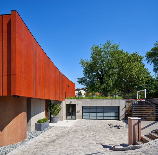 A contemporary residential exterior viewed from a paved courtyard or driveway. The building features a dramatic cantilevered upper volume clad in vertical rust-red metal panels, a concrete lower level with a wide glazed garage door, and timber-and-metal stair and balcony elements. Mature trees and a clear blue sky surround the home.