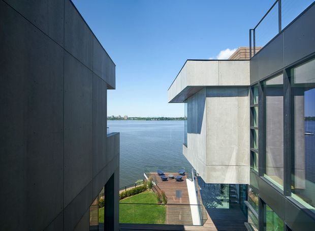 View between two contemporary waterfront building volumes, showing strong geometric concrete forms, extensive dark-framed glazing, a rooftop terrace or balcony, and a landscaped lawn with outdoor seating beside the water. The image is taken from an elevated exterior position and emphasizes the relationship between the architecture, terrace, and waterfront setting.