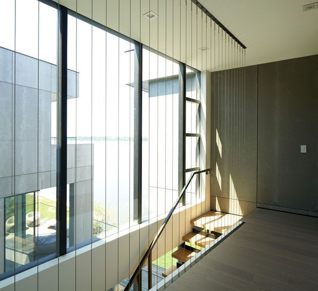 Contemporary interior landing or upper-level hallway overlooking a stairwell, with dramatic floor-to-ceiling glazing, slim dark window frames, a dark textured feature wall, and a linear suspended ceiling light. The open stair has a minimalist glass balustrade and timber-toned treads, while strong daylight creates a bright, refined atmosphere.