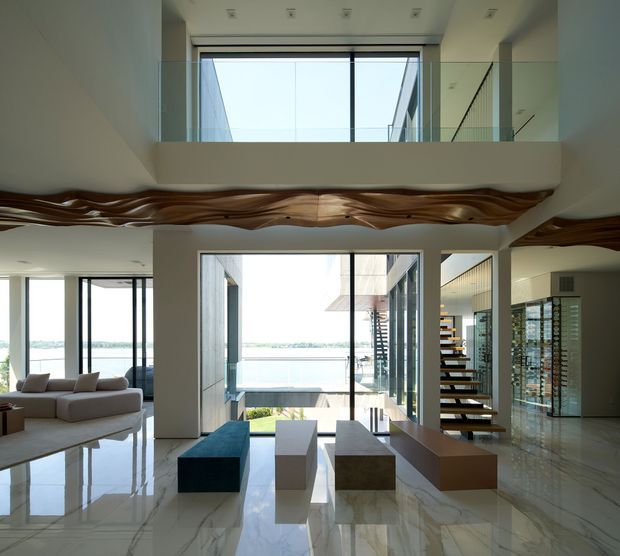 Bright, expansive double-height contemporary interior with polished pale stone flooring, extensive floor-to-ceiling glazing, a large sectional sofa, sculptural wood ceiling feature, floating open-riser staircase, and an upper-level glass balustrade. Water and landscape are visible beyond the glazing, while the main space reads as an open-plan living area with a strong architectural void and gallery-like proportions.