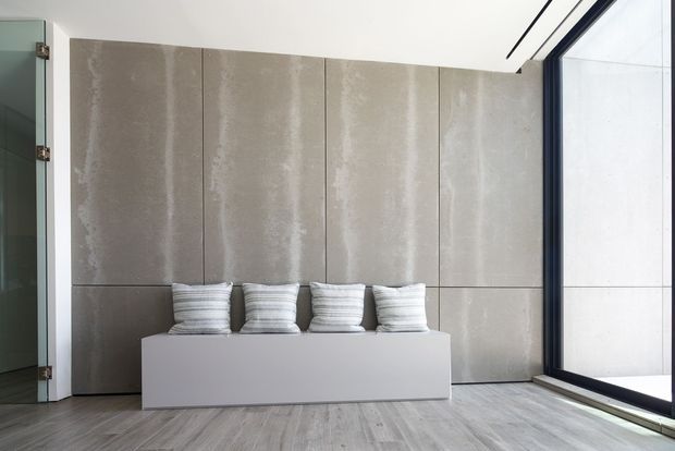 Minimalist interior featuring a long low white built-in bench with four striped cushions, set against a large-format vertically panelled grey concrete-look feature wall. Full-height dark-framed glazing occupies the right side, while pale timber-look flooring and a frameless glass door contribute to the clean, contemporary aesthetic.