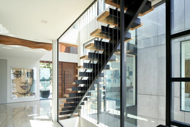 Bright contemporary interior dominated by a sculptural open-riser staircase with dark steel stringers, timber treads, and slender vertical balustrade wires. The stair rises beside full-height glazing and a pale stone or concrete feature wall, with polished light flooring, curved timber ceiling detailing, and an open living or entry area visible beyond.