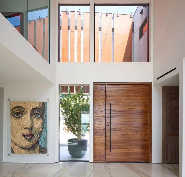Bright double-height residential entry foyer with a large timber pivot front door, polished light stone flooring, extensive upper-level glazing, and a sculptural stair opening at left. A large framed portrait hangs on the wall, while a potted tree is visible through the glazing. The architecture has clean modern lines and strong vertical window proportions, with warm timber contrasting against white walls and an orange-toned exterior courtyard facade.