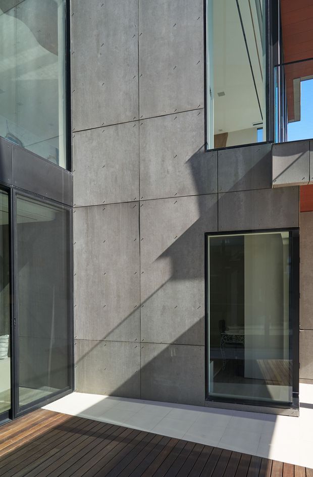 Modern multi-level building facade viewed from an outdoor deck or terrace. Large smooth grey concrete panel cladding with visible fixing marks forms the main wall, intersected by black-framed floor-to-ceiling glazing and projecting volumes. Warm timber soffits and dark timber decking contrast with the cool concrete, while strong sunlight creates geometric shadows across the facade.