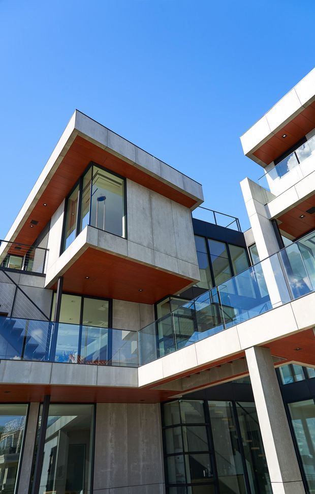 Upward-looking exterior view of a contemporary multi-level building with projecting concrete balconies and volumes, extensive dark-framed glazing, transparent glass balustrades, and warm timber-toned soffits. The composition emphasizes angular geometry, cantilevered forms, and a bright blue sky.