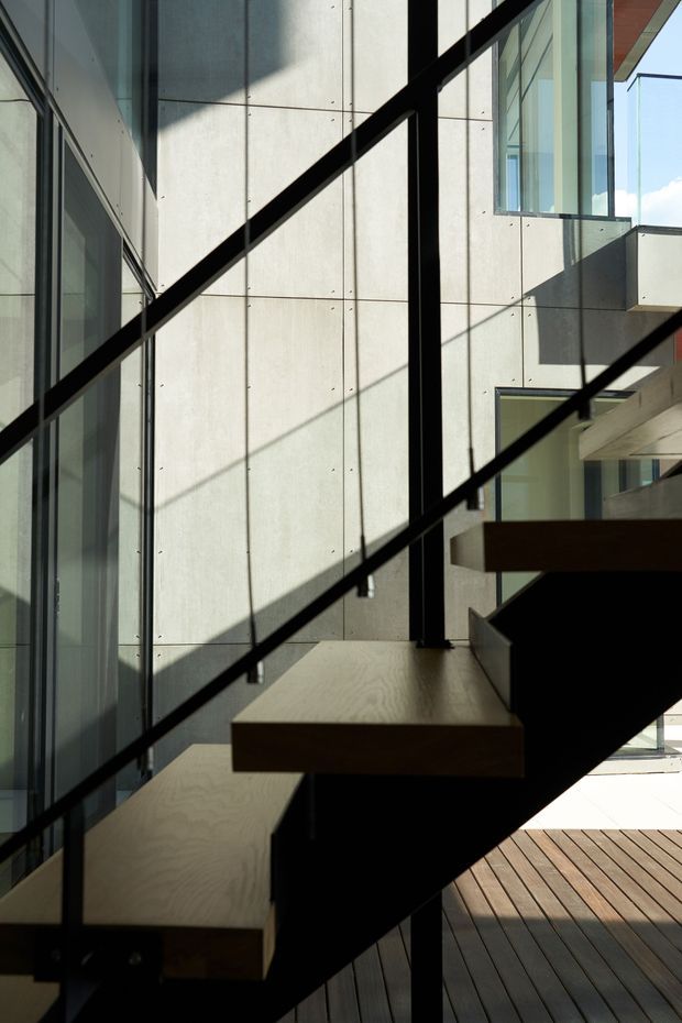 Modern minimalist interior view of a floating timber staircase with black metal stringers and slender black balustrade rails. The stair is set beside extensive floor-to-ceiling glazing and a pale exposed concrete panel wall, with a timber deck or flooring visible below and strong sunlight creating geometric shadows.