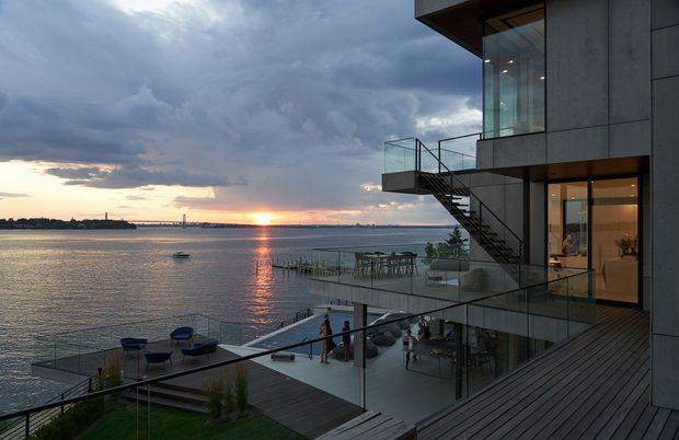 Contemporary multi-level waterfront home at sunset, featuring extensive glazing, dark vertical wall cladding, cantilevered terraces, glass balustrades, exterior stairs, and broad timber decking overlooking calm water. Outdoor dining and lounge areas are visible across the stepped terrace.
