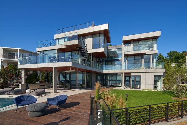 A large multi-level contemporary residence viewed from the landscaped outdoor area, featuring extensive floor-to-ceiling glazing, projecting cantilevered volumes, white and grey exterior surfaces, dark trim, glass balustrades, rooftop terraces, and exposed exterior stair elements. A timber deck, swimming pool, lawn, ornamental grasses, and outdoor seating occupy the foreground.