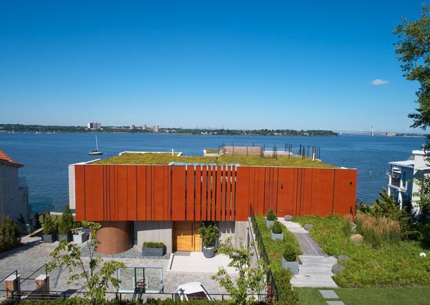 A contemporary waterfront residence viewed from above, featuring a bold rust-red vertically clad rectangular volume, a green roof, extensive glazing, landscaped terraces, timber decking, and a lower outdoor entertaining area. The home overlooks a broad body of water with boats and distant urban shoreline visible beyond.