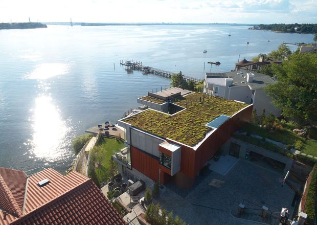 Aerial view of a contemporary waterfront residence beside a calm inlet, featuring a prominent green roof, flat-roofed forms, timber-toned cladding, terraces, and a private dock. The property is surrounded by neighbouring homes and landscaped outdoor areas.