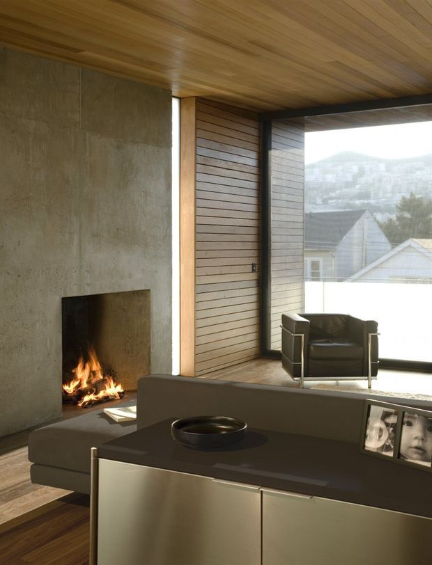 A refined modern living room with a built-in open fireplace set into a raw concrete wall, extensive warm timber panelling across the ceiling and feature wall, and large glazing overlooking a mountainous landscape. A low streamlined sofa and dark leather armchair sit beside a minimalist built-in sideboard, creating a warm, contemplative interior.