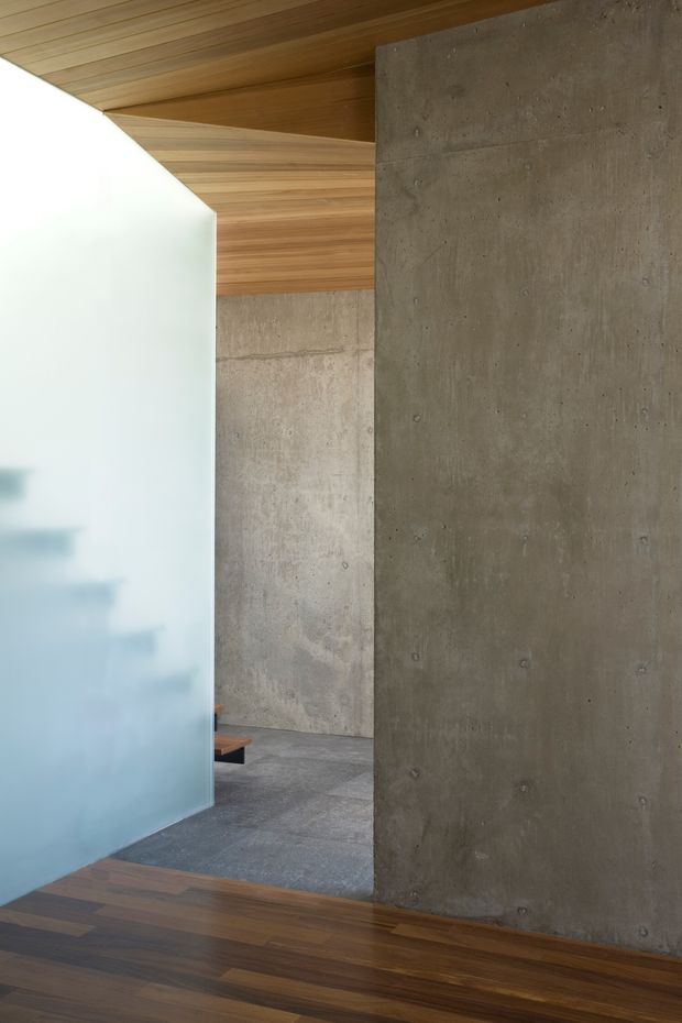 Interior architectural detail showing a minimalist circulation or living space with raw board-formed concrete walls and a warm timber-lined ceiling and floor. A translucent frosted glass partition partially screens the space, while a small section of seating is visible in the background. The composition emphasizes material contrast, clean geometric planes, and natural muted tones.