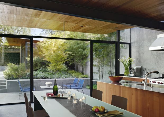 A refined open-plan dining and kitchen interior opening onto a landscaped courtyard through extensive black-framed glazing. The space features a long dining table, timber kitchen cabinetry with a stone benchtop, a stainless-steel cooktop and rangehood, a warm timber-lined ceiling, and a dark timber ceiling beam. The courtyard includes concrete retaining walls, paving, lush planting, and outdoor seating visible through the glass.