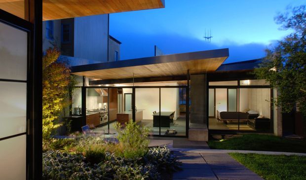 Twilight exterior view of a contemporary flat-roofed residence arranged around a landscaped courtyard. Extensive floor-to-ceiling glazing reveals illuminated interior living spaces, while broad projecting timber-lined roof canopies and dark slender frames create a strong horizontal composition. A paved path crosses the planted courtyard toward the house.