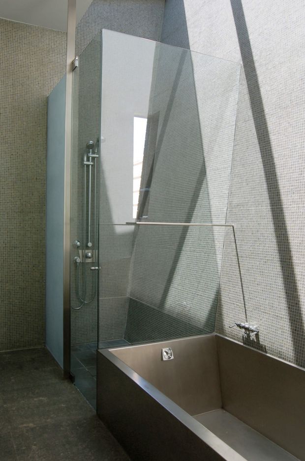 A tall, contemporary bathroom with a walk-in shower and deep rectangular bathtub. The walls are clad in small pale grey mosaic tiles, with a dark stone or concrete-look floor. A clear frameless glass shower screen, exposed shower fittings, and stainless-steel bath fixtures create a minimalist industrial feel. Strong natural light enters through a narrow vertical opening, casting dramatic geometric shadows across the tiled walls.