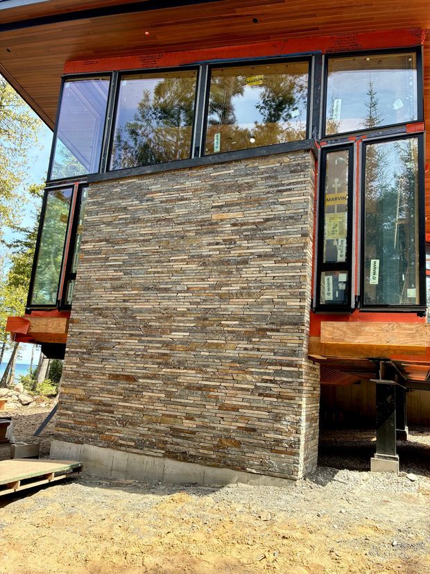 Exterior construction-stage view of a raised timber-clad building with extensive dark-framed glazing and a prominent stacked natural-stone wall. The structure sits above the ground on supports, with exposed timber soffits and unfinished ground works visible. Surrounding trees and a glimpse of water or sky appear through the glazing and around the building.
