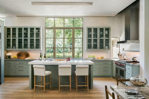 A bright, spacious traditional-style kitchen with a large central island and three upholstered bar stools. The room features muted sage-green cabinetry, glass-front upper display cabinets, pale stone countertops, warm timber flooring, a large timber-framed window, and stainless-steel cooking appliances with a substantial range hood. A dining table is partially visible in the foreground.