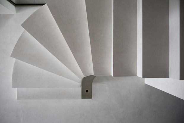 Overhead view of a sculptural modern stairway with broad white or light-grey concrete treads and angular geometry. The stair curves and fans around a central structural element, creating a minimalist, monochromatic composition with strong shadows and geometric rhythm.