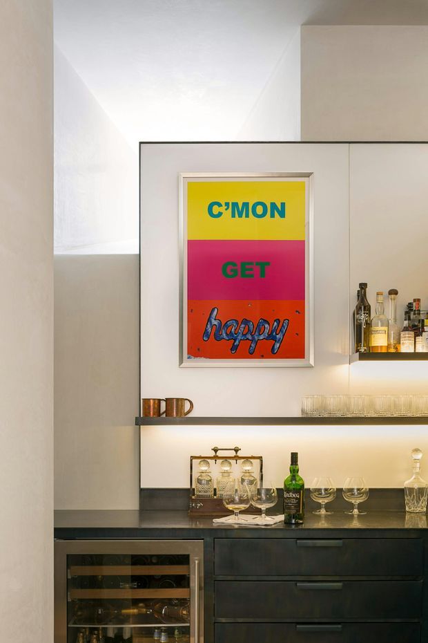 A compact built-in home bar with dark cabinetry, a black stone-like benchtop, illuminated glass shelving, bottle storage, wine glasses and spirits. A colourful framed typographic artwork provides a bold focal point against the otherwise neutral interior.