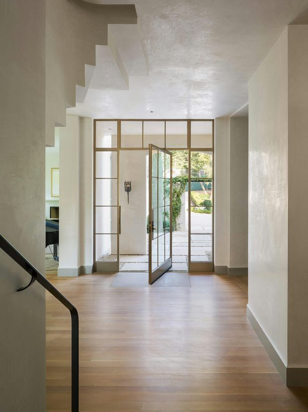 A bright, refined residential entry hall viewed from a stairway. The space has wide pale timber flooring, softly textured off-white plaster walls and ceiling, a black metal stair handrail, and a full-height glazed entrance with slim bronze or dark metal framing and a pivot door opening toward a landscaped courtyard. A glimpse of an adjacent room is visible to the left.
