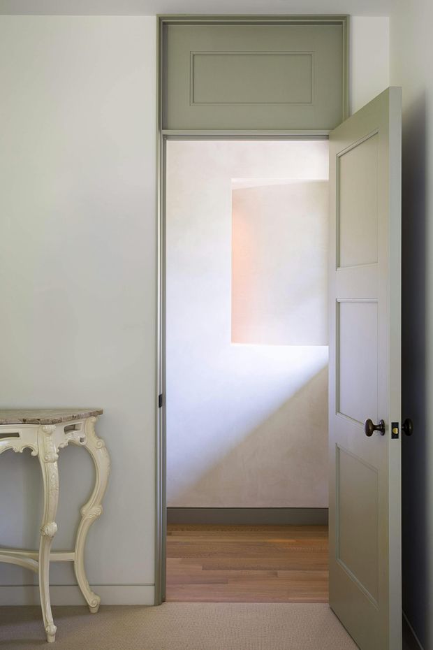 Interior view of a refined hallway or doorway with a partially open, panelled grey-green door and matching decorative transom panel. The space has pale painted walls with trim, a warm timber floor beyond the doorway, carpet in the foreground, and an ornate cream console table at the left edge. Soft natural light creates a calm, understated atmosphere.