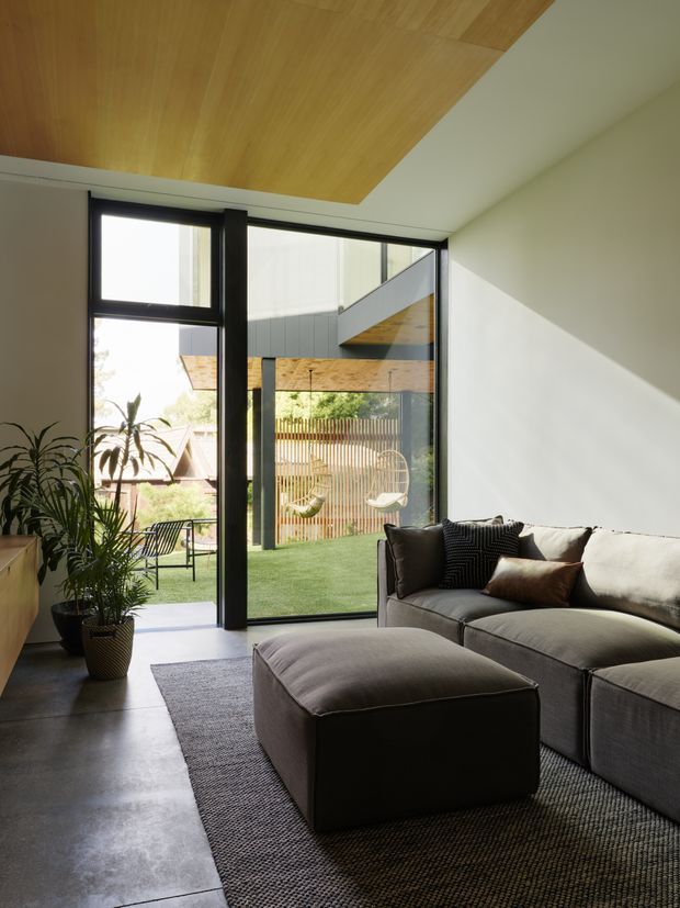 A bright contemporary living room with a large dark-framed glazed opening to a landscaped courtyard. The room features a low grey sectional sofa, oversized upholstered ottoman, textured area rug, potted plants, polished concrete flooring, white walls, and a warm timber-lined recessed ceiling. Exterior architectural elements and a small outdoor seating area are visible through the glazing.