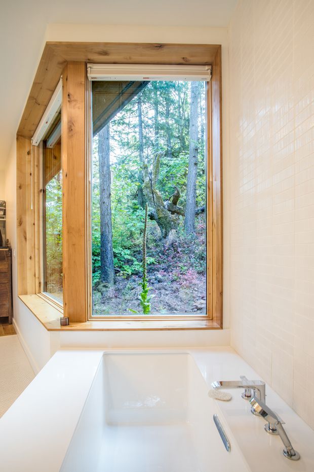 Bright bathroom featuring a freestanding white bathtub positioned beneath a large timber-framed window overlooking a dense forest. The room has white tiled walls, white fixtures, warm natural wood trim, and a chrome wall-mounted bath tap and hand shower. A partial mirror and shelving are visible at the left edge.