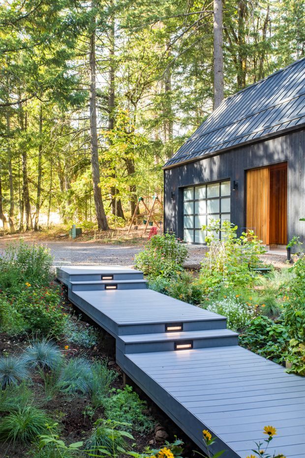 Exterior view of a compact dark-clad cabin or house set within a wooded garden. A broad grey decked walkway with integrated step lighting leads through landscaped planting toward the entrance. The building has a steep metal gable roof, dark vertical cladding, a large glazed garage-like opening, and a tall timber entry door.