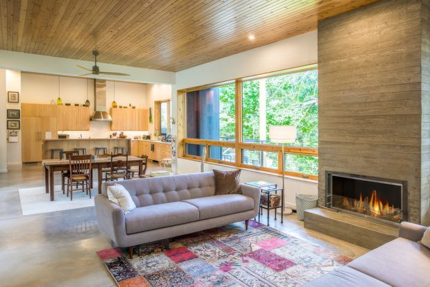Open-plan mid-century-inspired living room, dining area and kitchen with extensive timber ceiling panelling, warm timber cabinetry, polished concrete flooring, and large timber-framed windows overlooking greenery. A central concrete fireplace with a built-in gas fire anchors the living area, furnished with a grey tufted sofa, patterned rug, and light upholstered seating. The space is bright, warm, and connected to the outdoors.