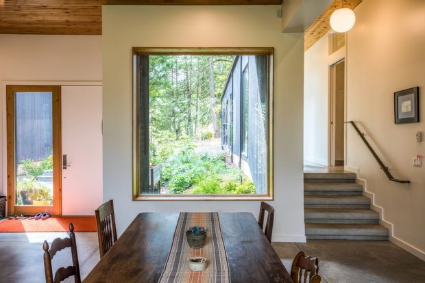 A warm, light-filled dining and entry area with a large timber-framed window overlooking a lush wooded garden. A substantial dark timber dining table occupies the foreground, while a staircase with grey tiled or stone treads rises along the right wall. The space features white plaster walls, exposed timber ceiling boards, timber detailing, simple framed artwork, and a glazed entry door at left.
