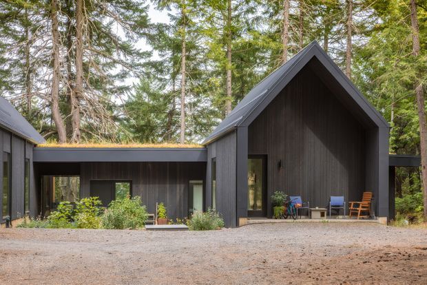 A dark-clad contemporary cabin-style residence set within a forested site. The composition features a steep gabled central volume connected to lower flat-roofed wings, with large openings, a covered entry or outdoor sitting area, and a gravel forecourt. Vertical dark timber cladding and a subtly planted roof on the lower connection create a restrained, natural woodland aesthetic.