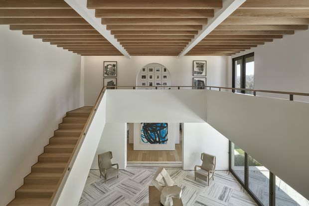 Double-height interior landing overlooking a bright living area, with a floating timber staircase, exposed timber ceiling battens, white walls, extensive glazing, and curated artwork. The lower level includes lounge seating and a sculptural central table on a patterned rug.