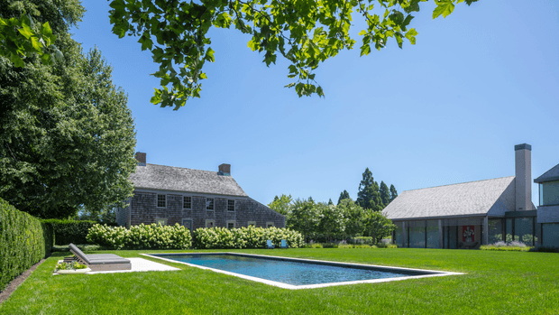 A landscaped residential courtyard viewed from the lawn, featuring a long rectangular swimming pool with a white coping edge, manicured hedges, mature trees, and two connected shingle-clad pitched-roof buildings with tall chimneys in the background. The scene has a refined traditional rural character under clear blue skies.
