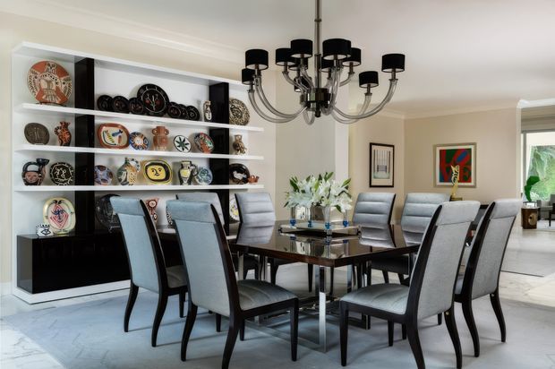 A refined formal dining room with a large rectangular glass-topped dining table surrounded by upholstered high-back chairs. A dramatic black multi-arm chandelier hangs above the table, while a built-in white shelving unit with dark lower cabinetry displays decorative plates and objects along the left wall. Framed artwork, a floral arrangement, neutral walls, pale flooring, and abundant daylight create a polished, elegant interior.