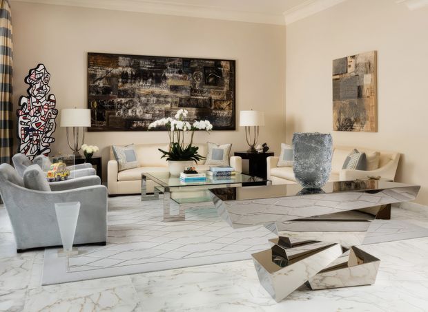 A sophisticated formal living room with a neutral cream and grey palette. The room features two light sofas, plush grey armchairs, a glass coffee table with a metallic base, sculptural metallic and marble-look accent tables, abstract artwork, table lamps, orchids, and a large pale geometric rug over polished white flooring. Traditional wall moulding and cornicing frame the otherwise contemporary, art-focused interior.