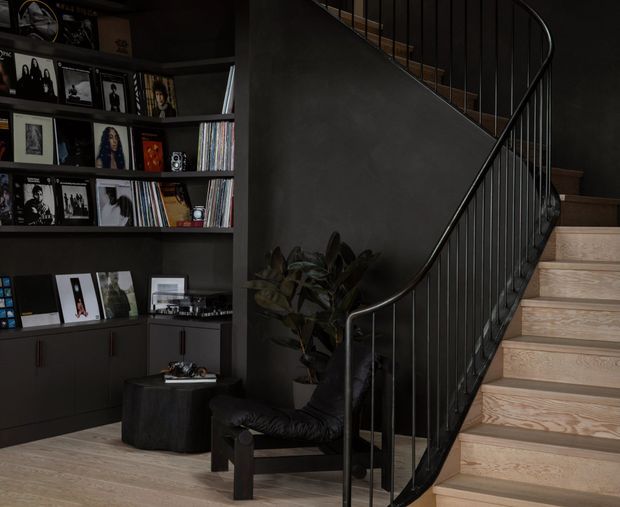 A moody, refined interior stairway with pale timber stairs and flooring, dark charcoal walls, and a slim black metal balustrade that curves around the stair. A built-in dark shelving and cabinetry wall displays records, framed artwork, and books, while a black leather lounge chair and indoor plant occupy the landing area.