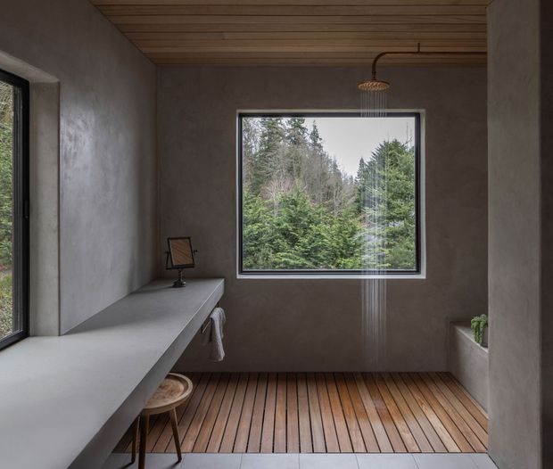 A minimalist bathroom with a long floating concrete vanity along the left wall, a wall-mounted basin area, timber slatted ceiling, and a warm timber floor with linear drainage near the foreground. A large picture window frames dense forest greenery, while a small raised planter or ledge sits at the right. The room has raw textured plaster walls, restrained detailing, and a calm, natural atmosphere.