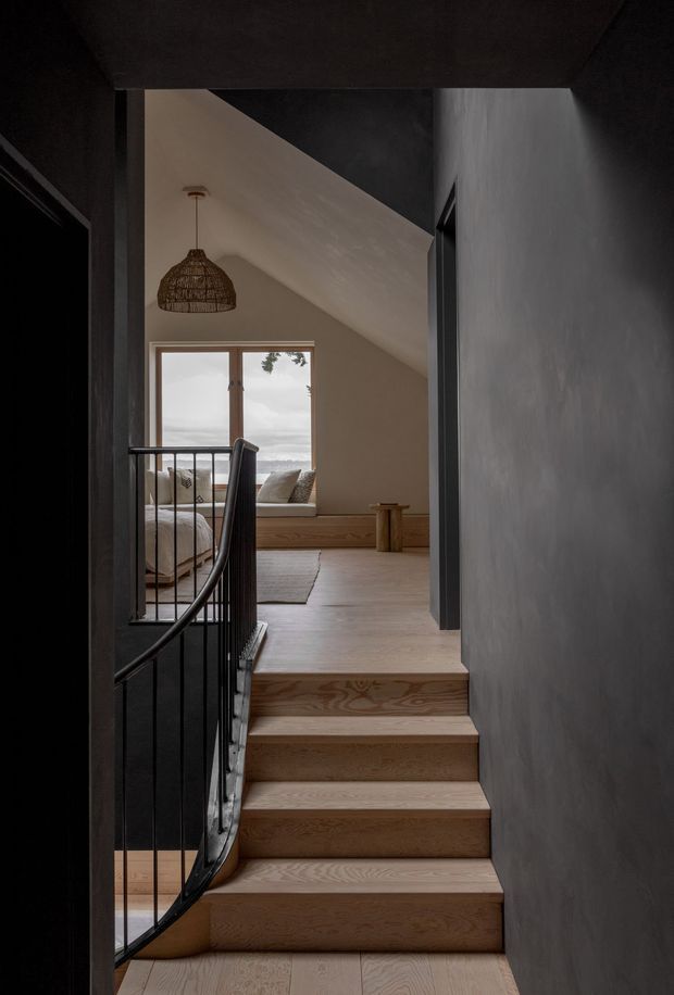 A moody interior view up a short timber staircase toward a loft-like bedroom or sitting area. The stair has pale timber treads and risers with a slender black metal balustrade, while dark charcoal walls and trim frame the space. At the upper level, a low built-in window seat with cushions sits beneath a sloping ceiling, and a woven pendant light hangs above. A large window provides a muted outlook, adding soft natural light to the otherwise warm, subdued interior.