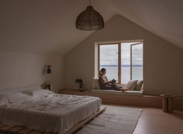 A warm, softly lit bedroom with a pitched ceiling and a large timber-framed window seat overlooking a body of water. The room features a low platform bed with white linen, pale timber flooring and built-in window-seat joinery, a woven pendant light, a small side table, and restrained natural decor. A person is seated in the window alcove, reading or relaxing.