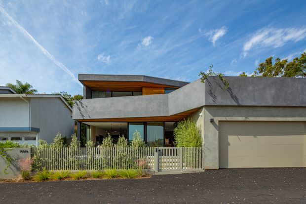 Street-facing view of a contemporary two-storey residence with strong horizontal concrete forms, a projecting upper-level balcony, warm timber soffit, extensive glazing, and an integrated double garage. A metal picket fence and landscaped planting sit in front of the house.