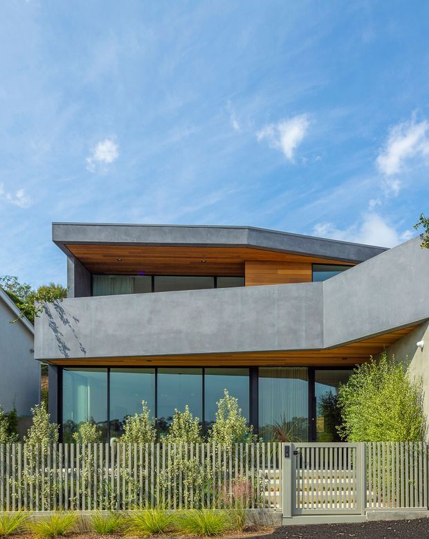 Modern two-storey residential facade with a strong horizontal composition, broad concrete balcony walls, extensive dark-framed glazing, and warm timber soffits and upper-level cladding. The house is screened by a vertical metal front fence and landscaped planting, with a partly cantilevered flat roof and clean geometric forms.