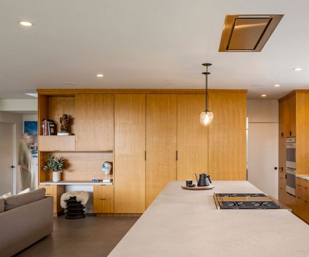 Warm contemporary open-plan kitchen with a large pale stone island in the foreground, integrated gas cooktop, timber cabinetry, and a central pendant light. Full-height natural timber joinery includes concealed storage and open display shelving, while a living area with a sofa is visible to the left. Recessed ceiling lights and a ceiling ventilation unit complete the clean, refined interior.