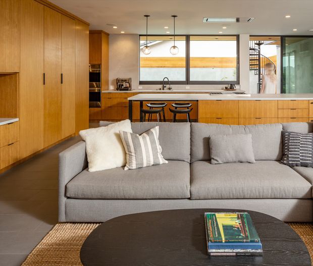 A warm, open-plan contemporary interior viewed from the living area toward a timber-lined kitchen. A large grey sectional sofa and round dark coffee table occupy the foreground, with a timber kitchen, island bench, pendant lights, and stools behind. Full-height glazing and an internal stair are visible in the background, along with neutral finishes and integrated cabinetry.