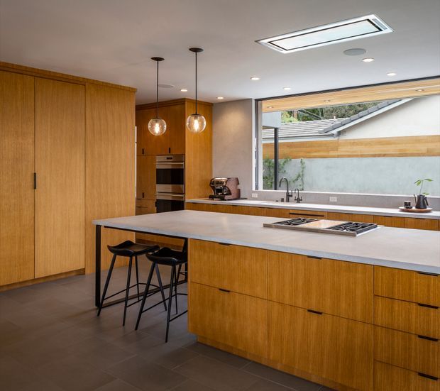 A warm contemporary kitchen dominated by full-height and island cabinetry in a honey-toned timber veneer. The large island has a pale stone benchtop, integrated cooktop and two black bar stools. Pendant lights hang above the island, while a wall of cabinetry includes built-in ovens. Dark tiled flooring, a neutral ceiling, recessed lighting and a large glazed opening to an exterior courtyard complete the space.