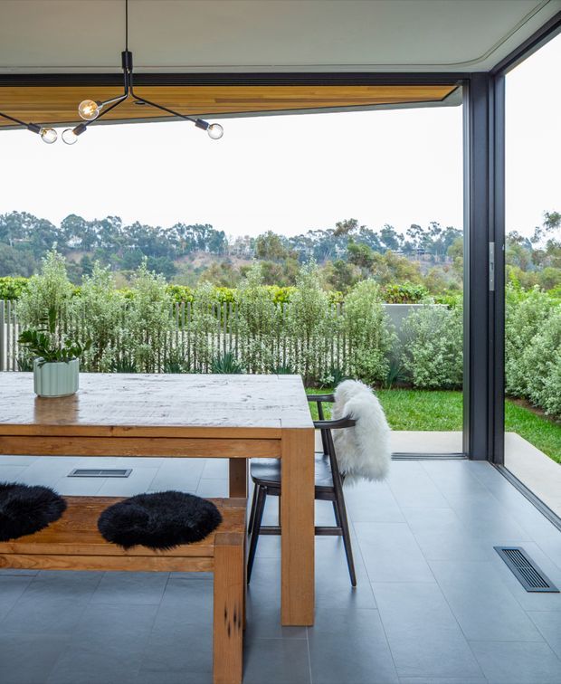 A bright contemporary dining area opening directly onto a landscaped garden through expansive dark-framed glazing. The space features a substantial timber dining table with a white stone top, matching timber bench, black chairs with sheepskin throws, a sculptural pendant light, pale large-format tiled flooring, and a timber-lined ceiling detail. Dense hedging and a green garden are visible beyond the glass.
