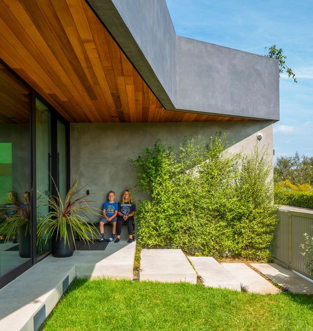 Exterior view of a contemporary home entry and covered porch. The facade combines smooth grey rendered walls with a dramatic projecting flat roof and warm timber-lined soffit. Full-height dark-framed glazing opens onto a small concrete porch, with concrete stepping slabs, lawn, shrubs, and a metal balustrade defining the landscaped approach.