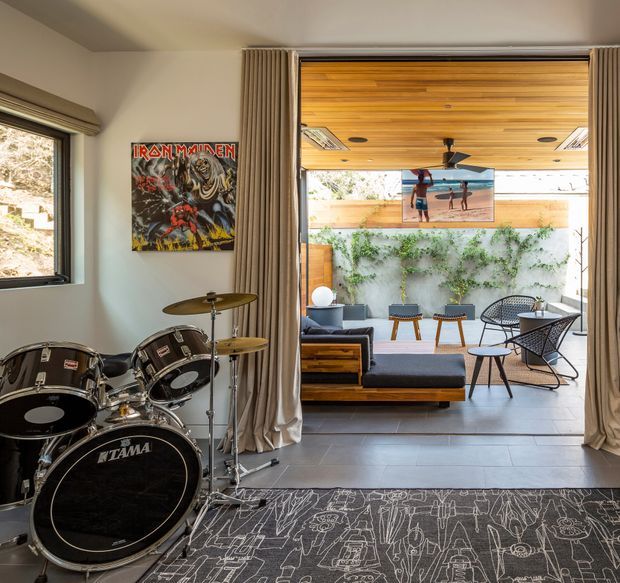 Interior view of a music-oriented living space with a full drum kit in the foreground, opening through floor-to-ceiling sliding glazing to a covered outdoor lounge. The room features pale walls, full-height beige curtains, grey tiled flooring, and a dark patterned rug. Outside, a timber-lined ceiling, concrete feature wall with climbing greenery, and mid-century-inspired outdoor seating create a bright indoor–outdoor entertaining area.